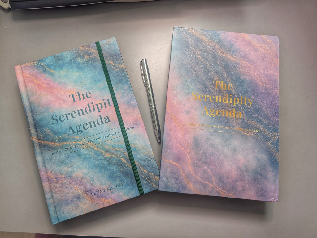 The Serendipity Agenda with its sleeve case on a desk, by Penny Naoum, featuring a pastel marbled cover with blue, pink, and purple tones and gold foil text, displayed on a gray surface alongside a silver branded pen.