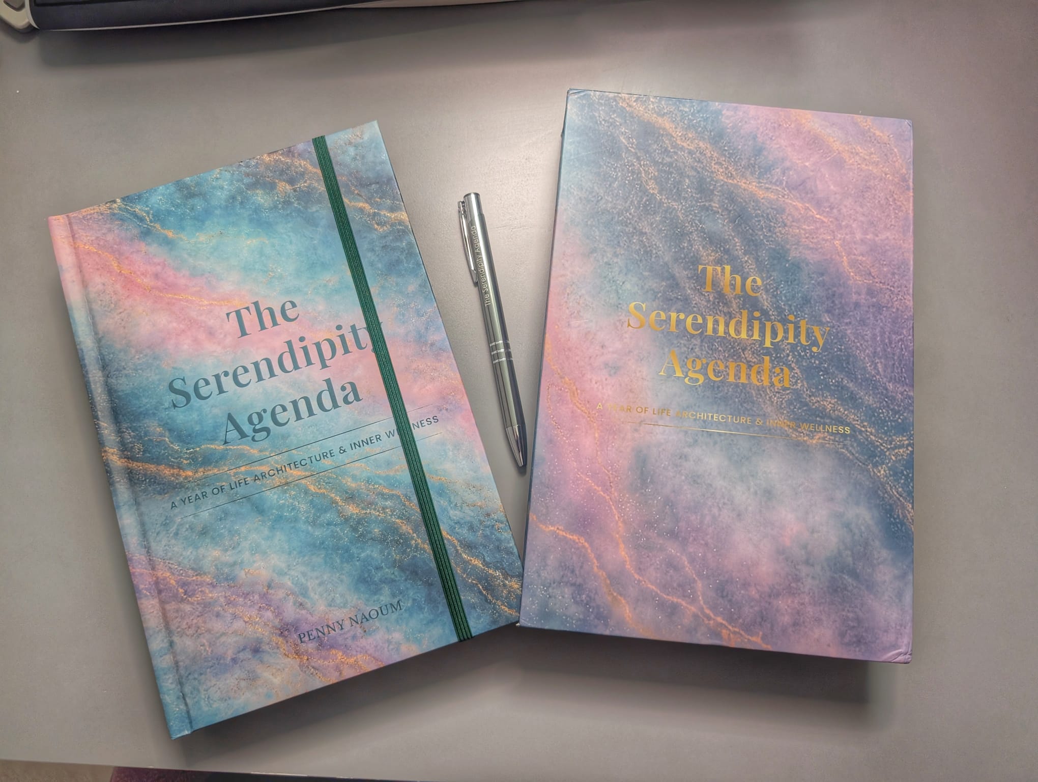 The Serendipity Agenda with its sleeve case on a desk, by Penny Naoum, featuring a pastel marbled cover with blue, pink, and purple tones and gold foil text, displayed on a gray surface alongside a silver branded pen.