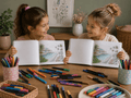 Two little girls holding open books with coloring pages of a pool, surrounded by colorful markers and crayons on a wooden table.