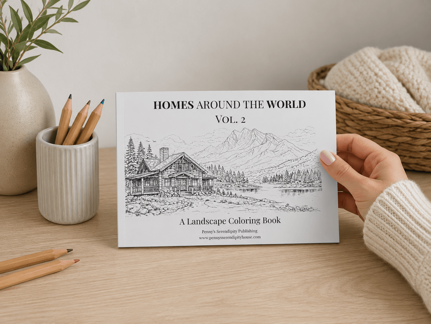 Coloring book titled 'Homes Around the World Vol. 2' held by a hand on a wooden surface with stationery items.