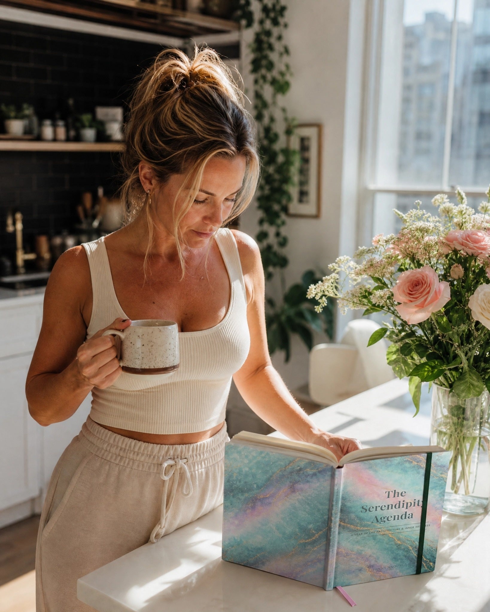 Woman in a kitchen holding a mug and reading The Serendipity Agenda book with a flower arrangement on the counter.
